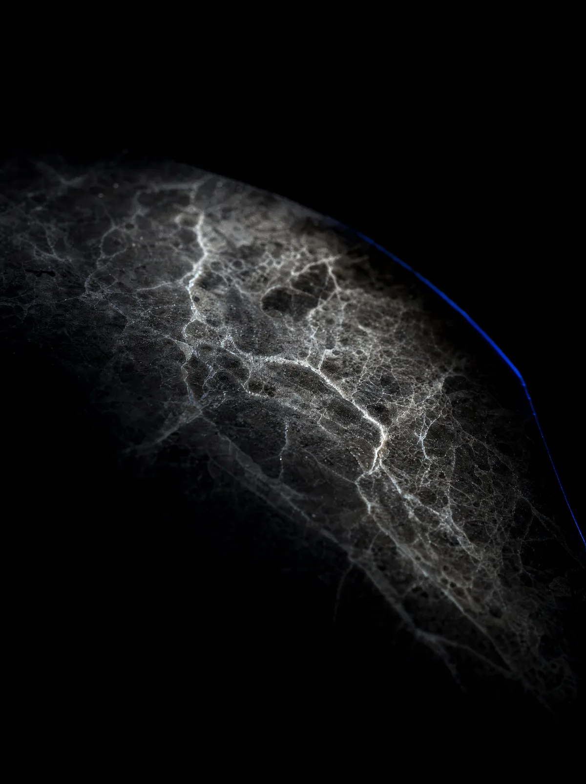 A macro photograph of grey and charcoal veined marble lit by a raking light, emerging from deep shadow